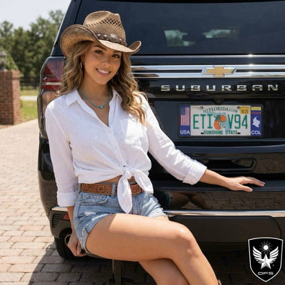 A woman wearing a straw cowboy hat, white button-up shirt tied at the waist, and denim shorts sits in front of a black Chevrolet Suburban upgraded with DFS Colombia Patented License Frame Official Gift.